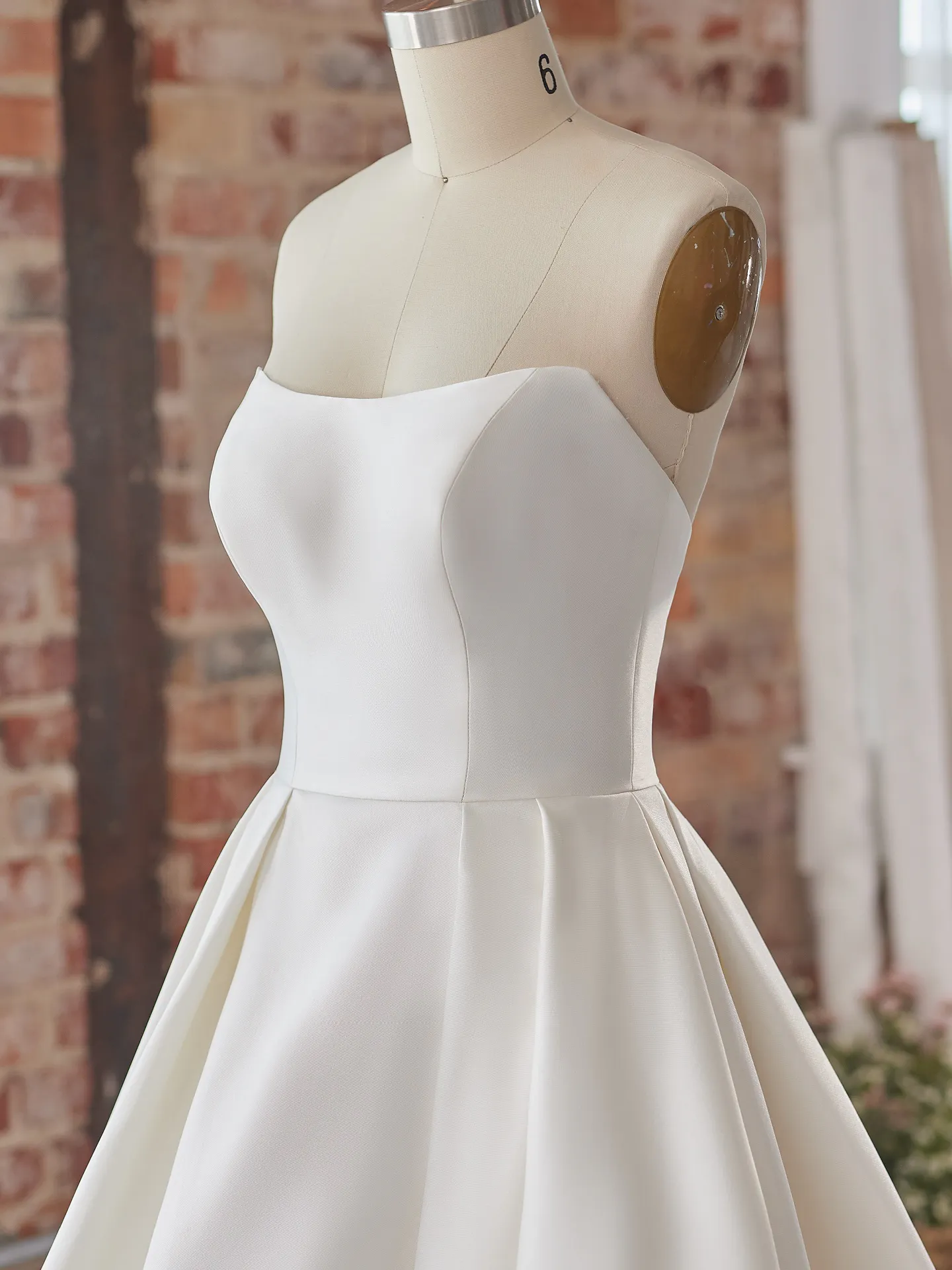 Maggie Sottero Selena satin A-line gown on mannequin, brick wall background.