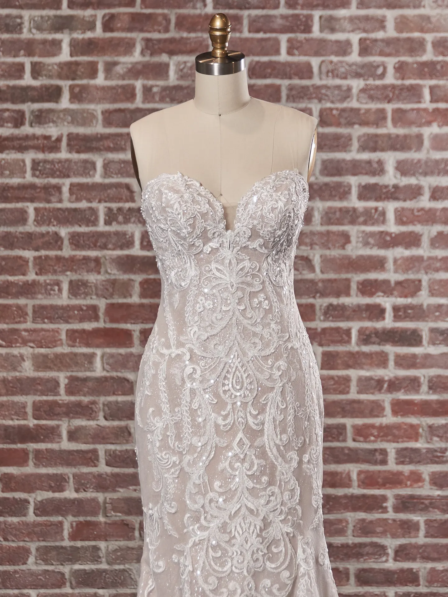 Maggie Sottero Frederique lace gown on mannequin, front view against brick wall background.