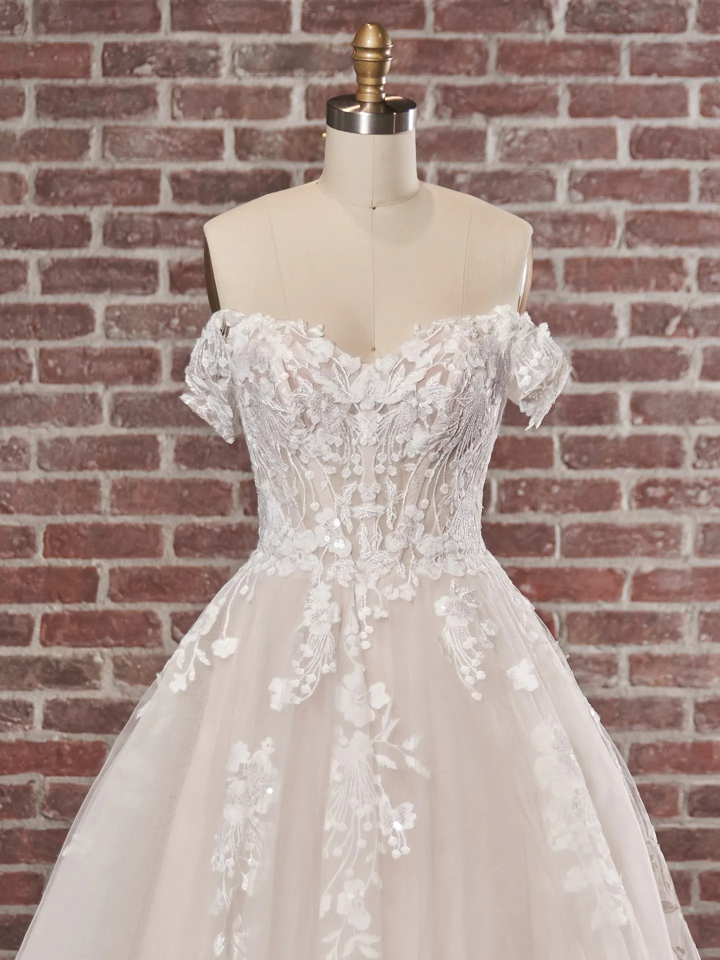 Maggie Sottero Harlem off-shoulder gown front view, floral lace bodice, brick wall backdrop