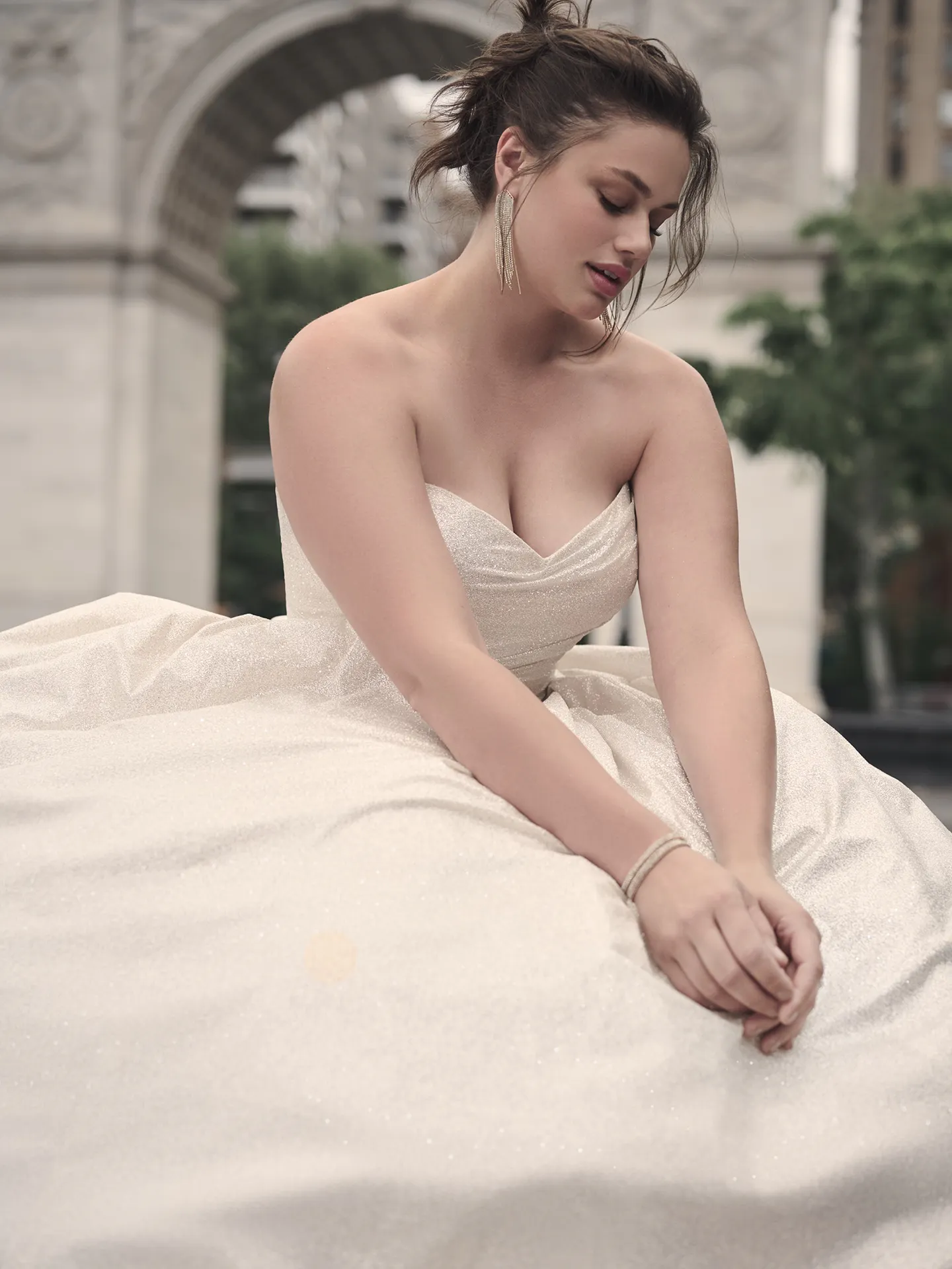 Anniston ballgown with shimmery fabric by Maggie Sottero, model seated outdoors.