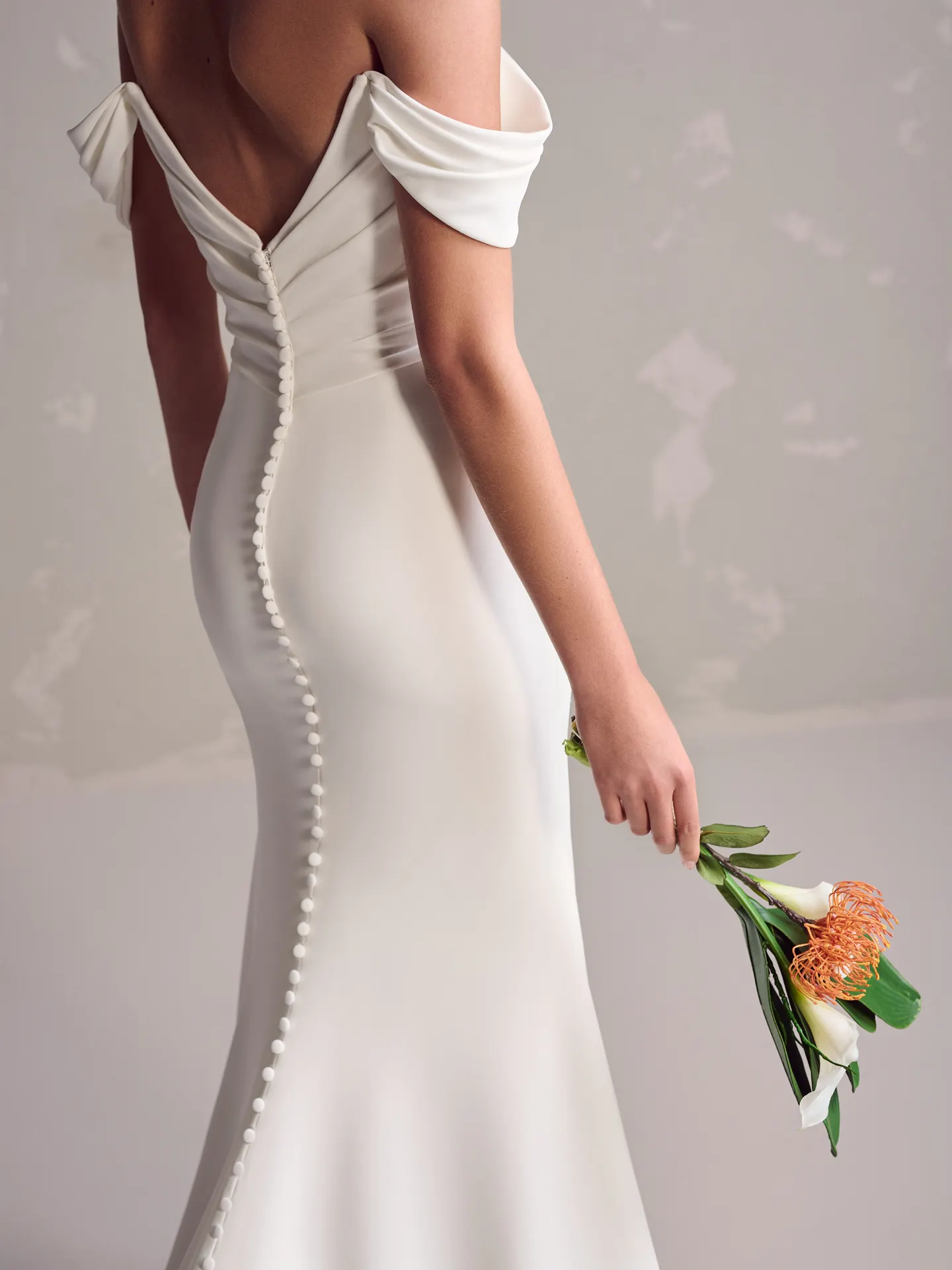 Maggie Sottero Summer back view with buttons, off-shoulder crepe dress, model holds flowers.