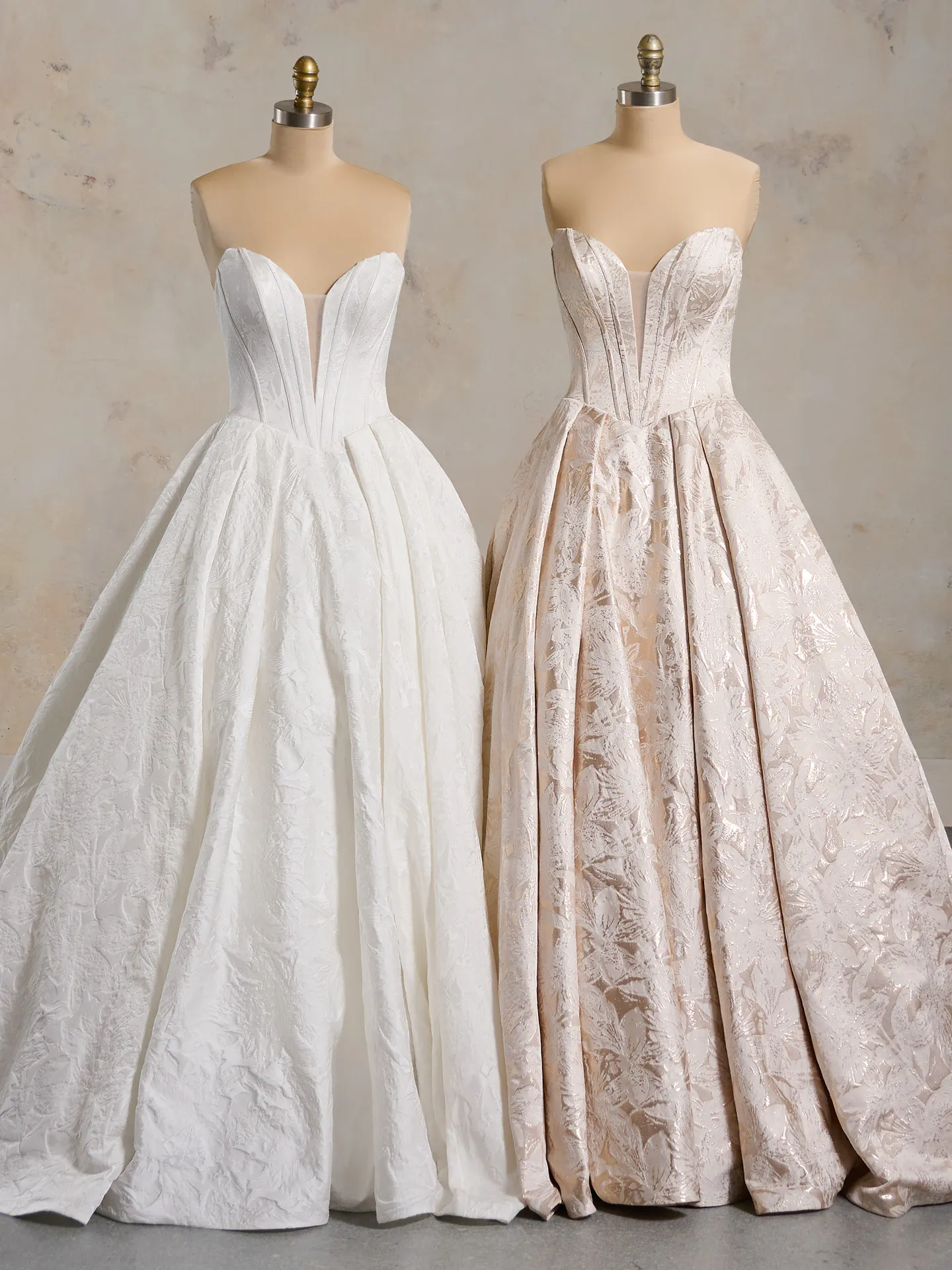 Two strapless floral ball gown wedding dresses on mannequins, one white, one blush.