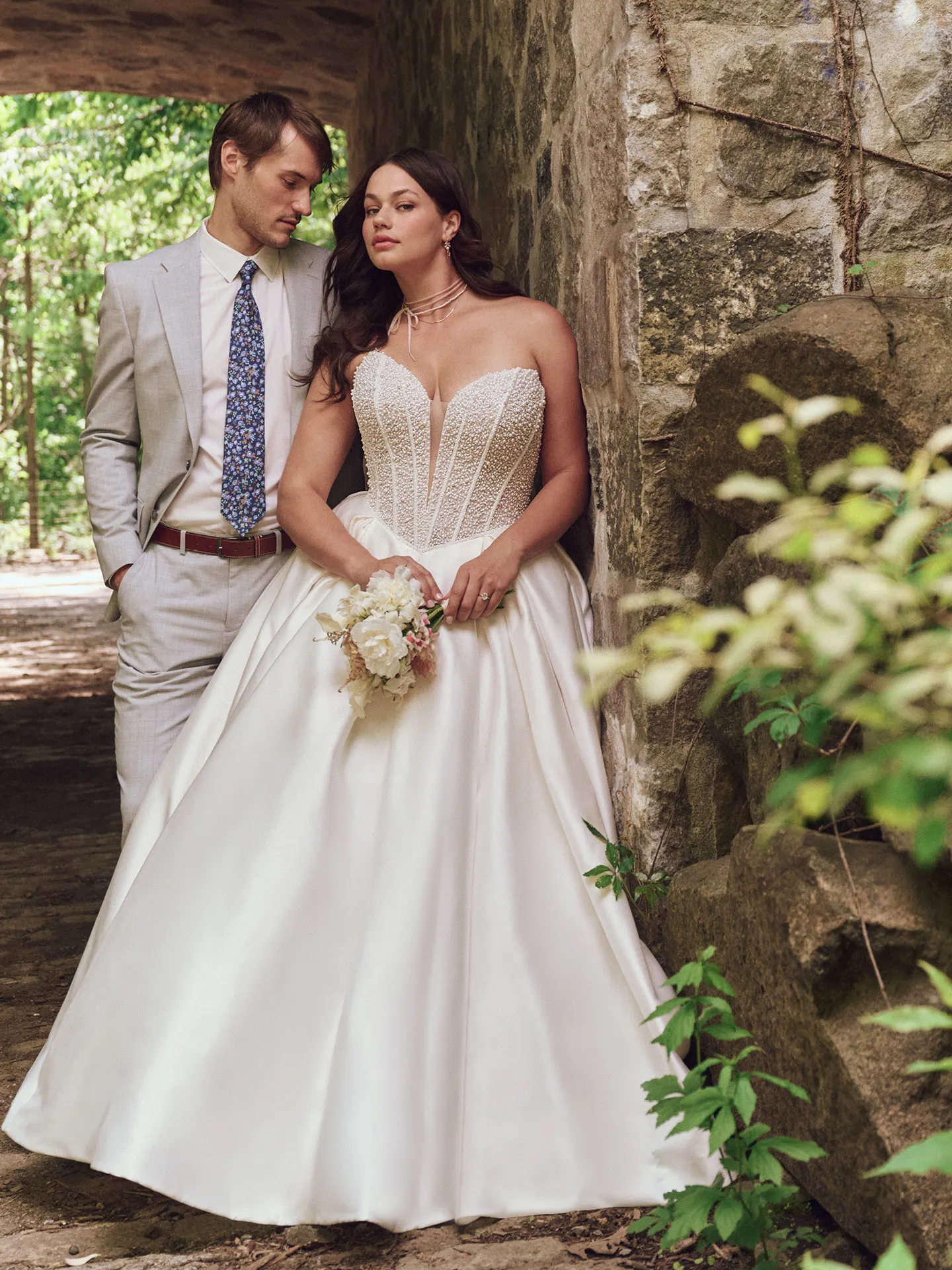 Maggie Sottero Tina front view, pearl basque waist ball gown amidst stone and greenery.
