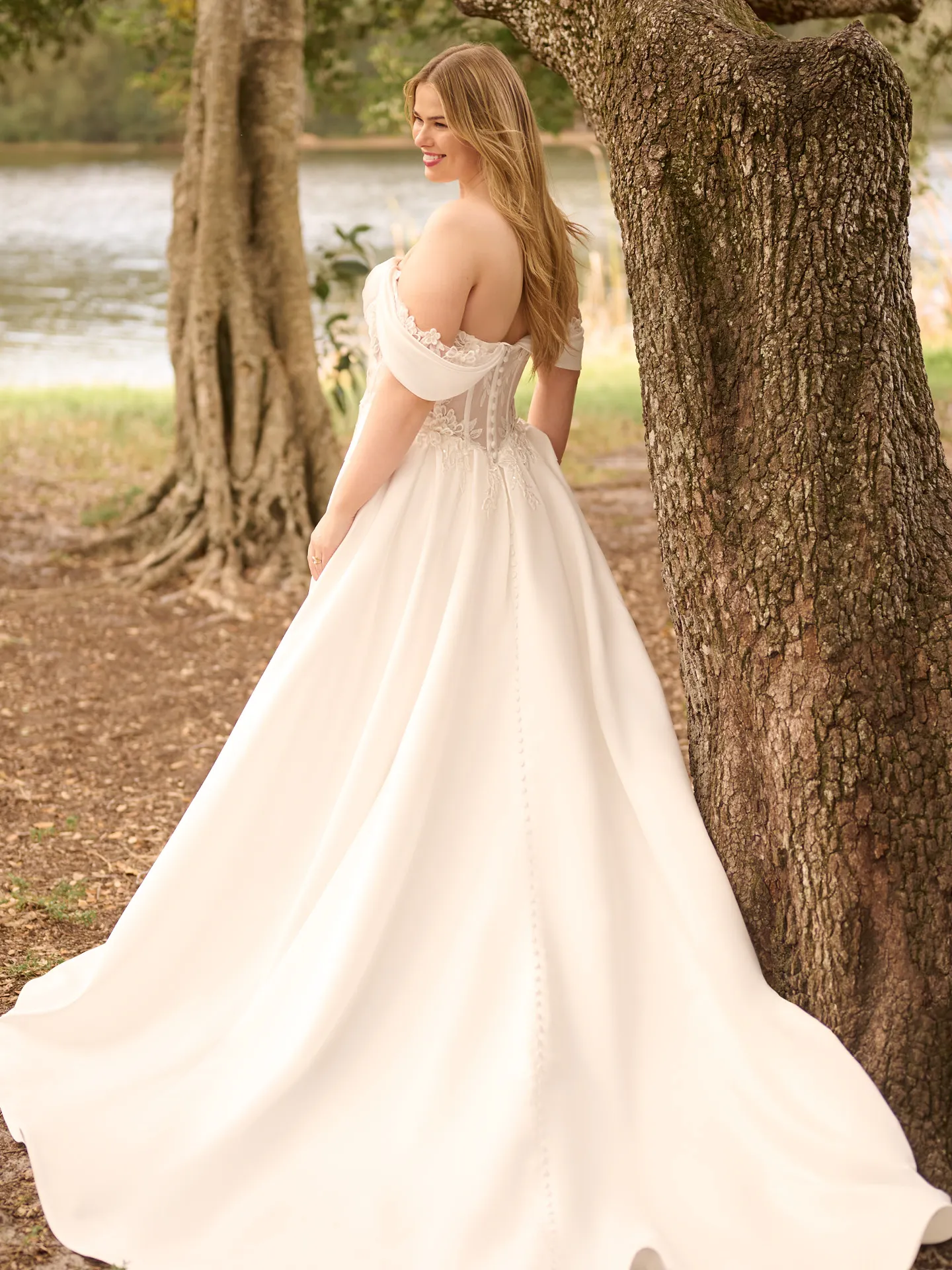 Rebecca Ingram Sable back view; floral peekaboo sweetheart A-line wedding gown near trees and lake.