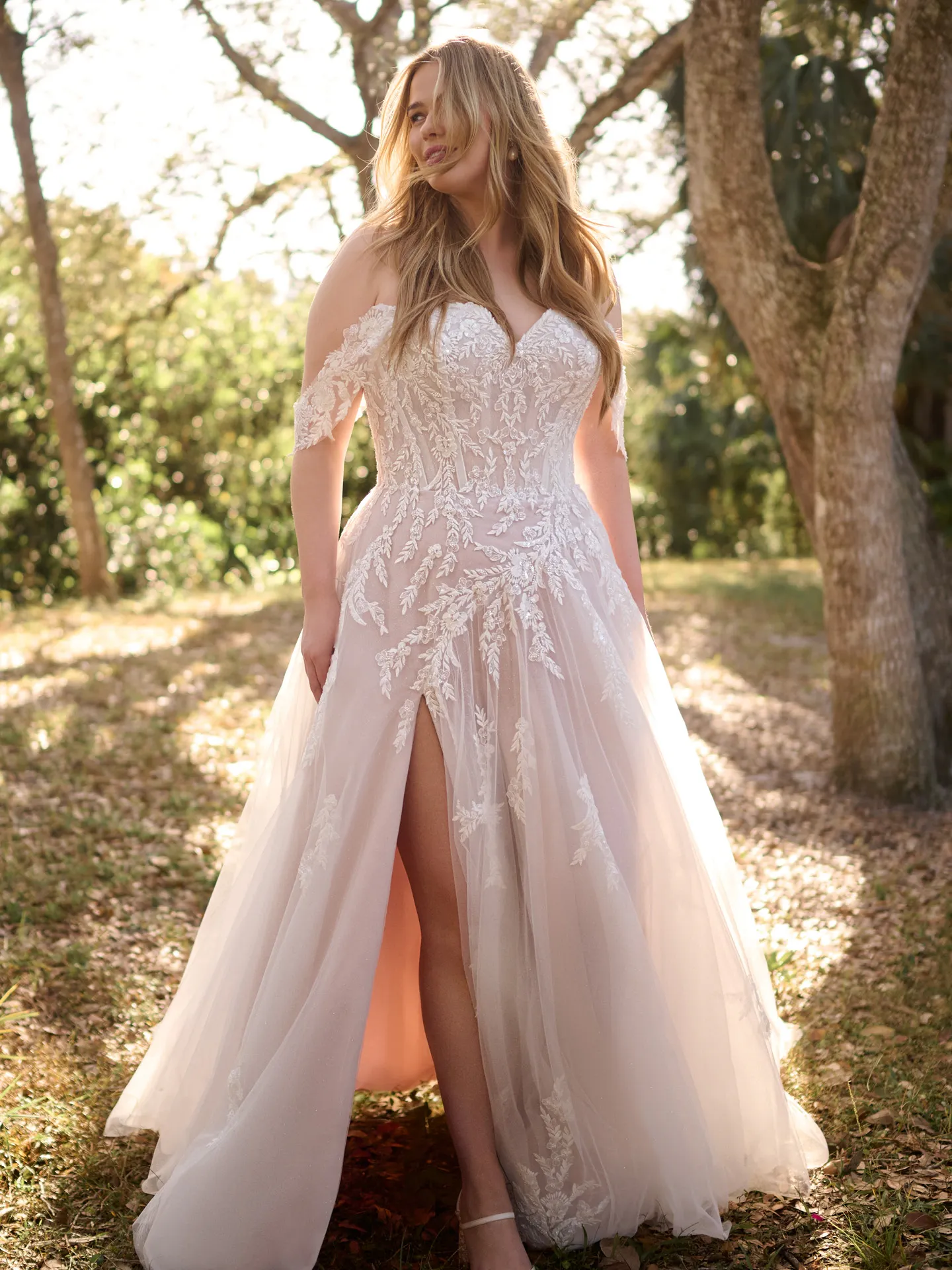 Rebecca Ingram Victoria floral slit A-line gown front view in a sunlit garden setting.