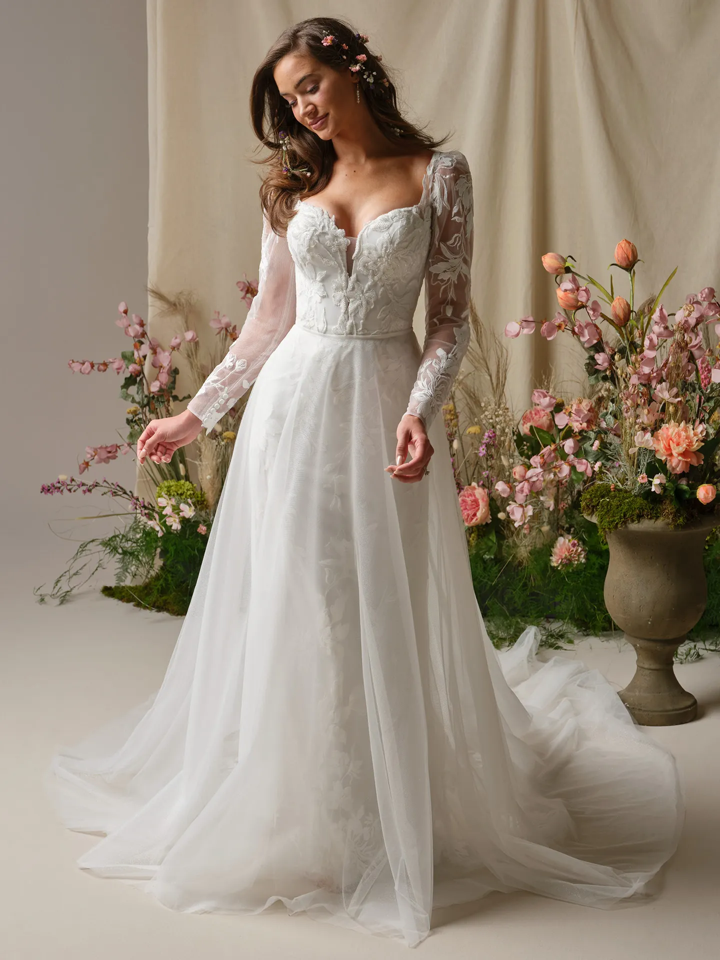 Maggie Sottero wedding dress, front view with lace sleeves and floral backdrop.